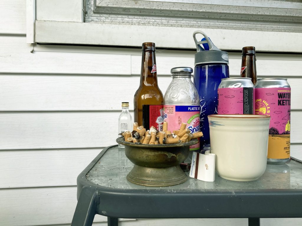 Empty bottles, cans, water bottles, and a full bowl of used cigarettes sitting on a table