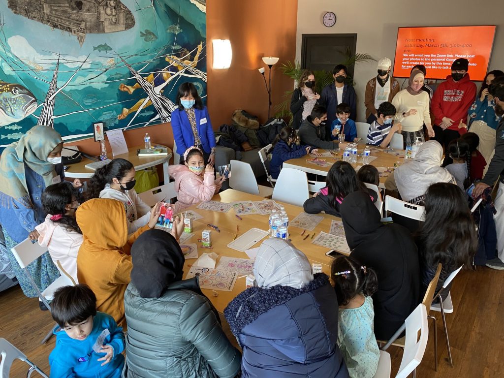 Workshop participants wearing face masks sitting and standing around 2 tables with colored drawings and crayons