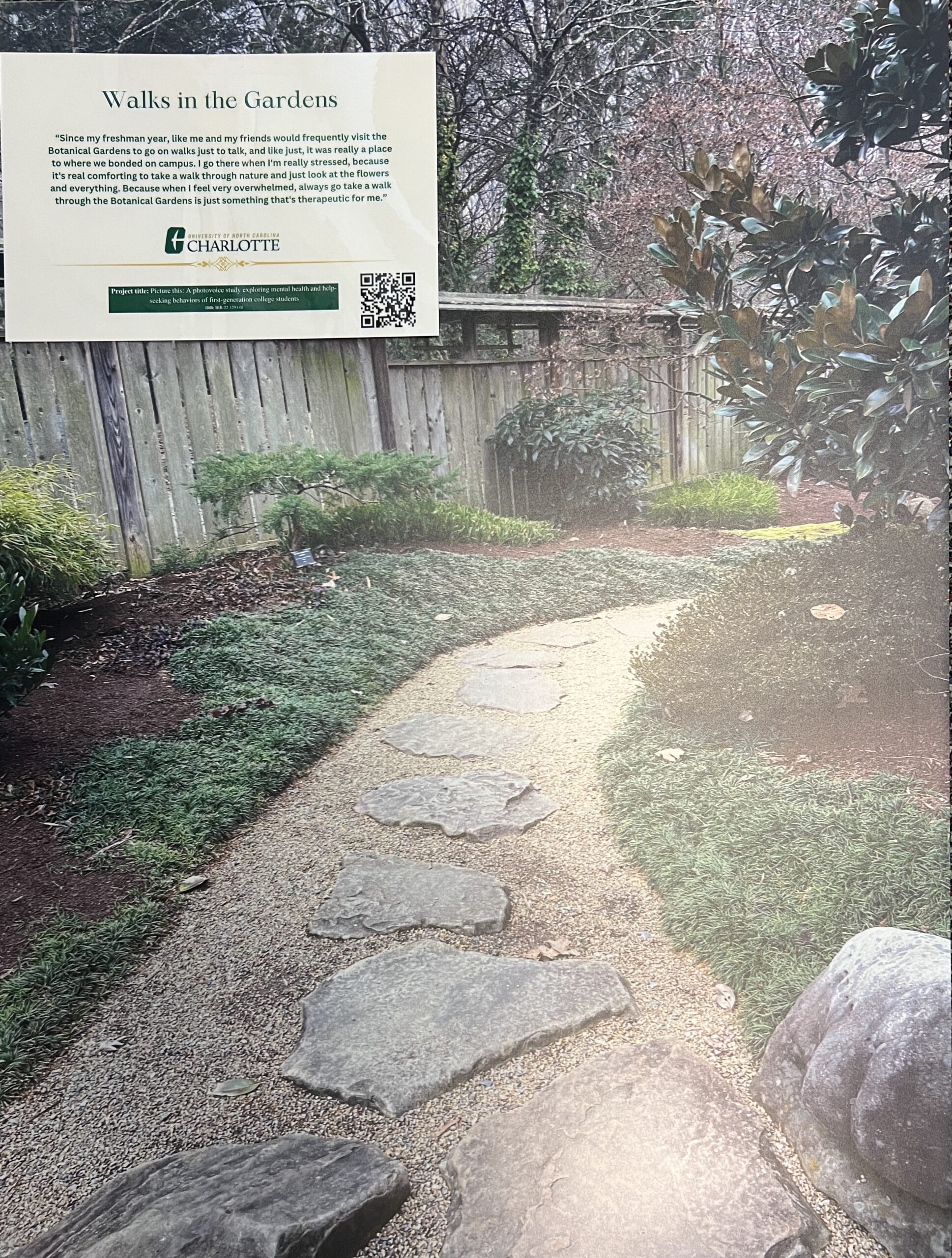 A path through a garden next to a fence, with a sign in the upper left hand corner.  
