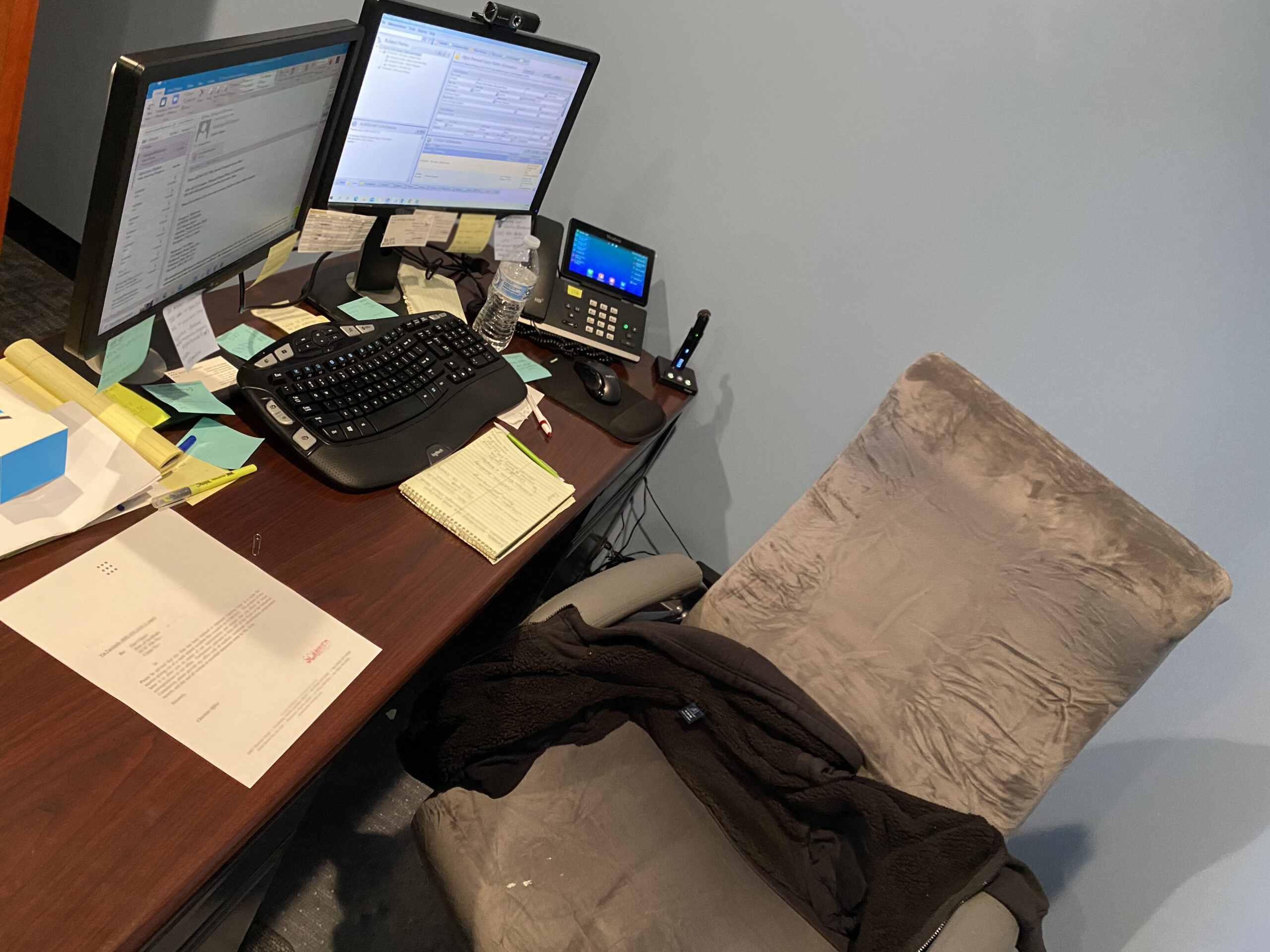 Desk with 2 computers, papers and a brown chair pulled out from the desk. 