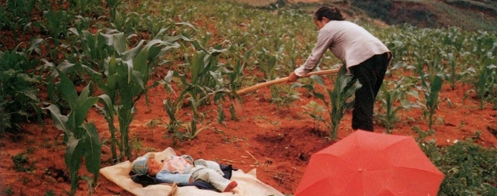 Woman working in a field of crops while her baby lays on a blanket on the ground close by.